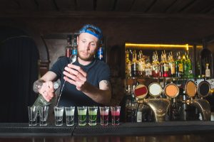 A barman with a beard makes colored alcoholic shots at the bar. Creation of showrooms at the bar restaurant