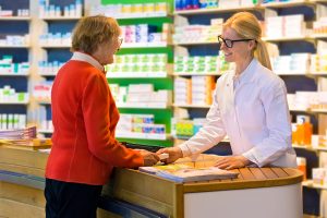 Happy senior citizen customer in red standing at pharmacy counter as pharmacist in eyeglasses and lab coat hands her a medication order.