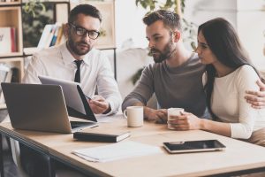 I need your signature here. Confident young man in shirt and tie holding some document and pointing it while sitting together with young couple at the desk in office